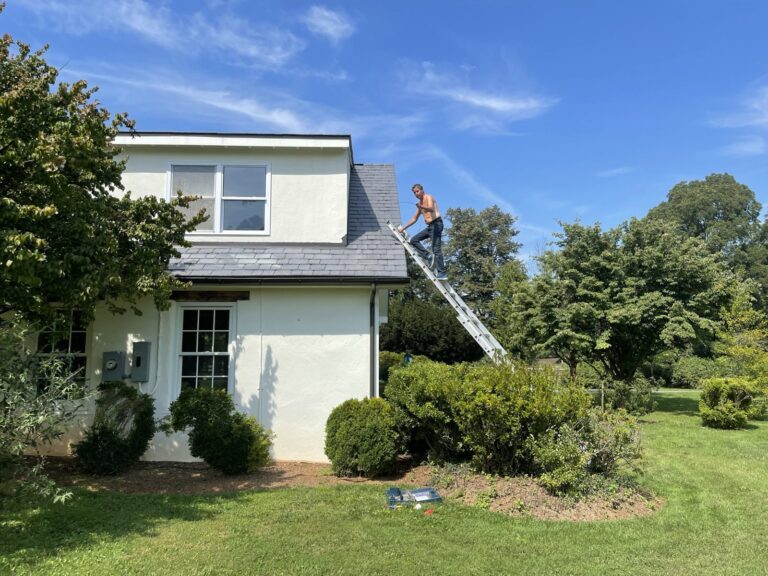 Installer on a ladder working on the roofline of a white farmhouse